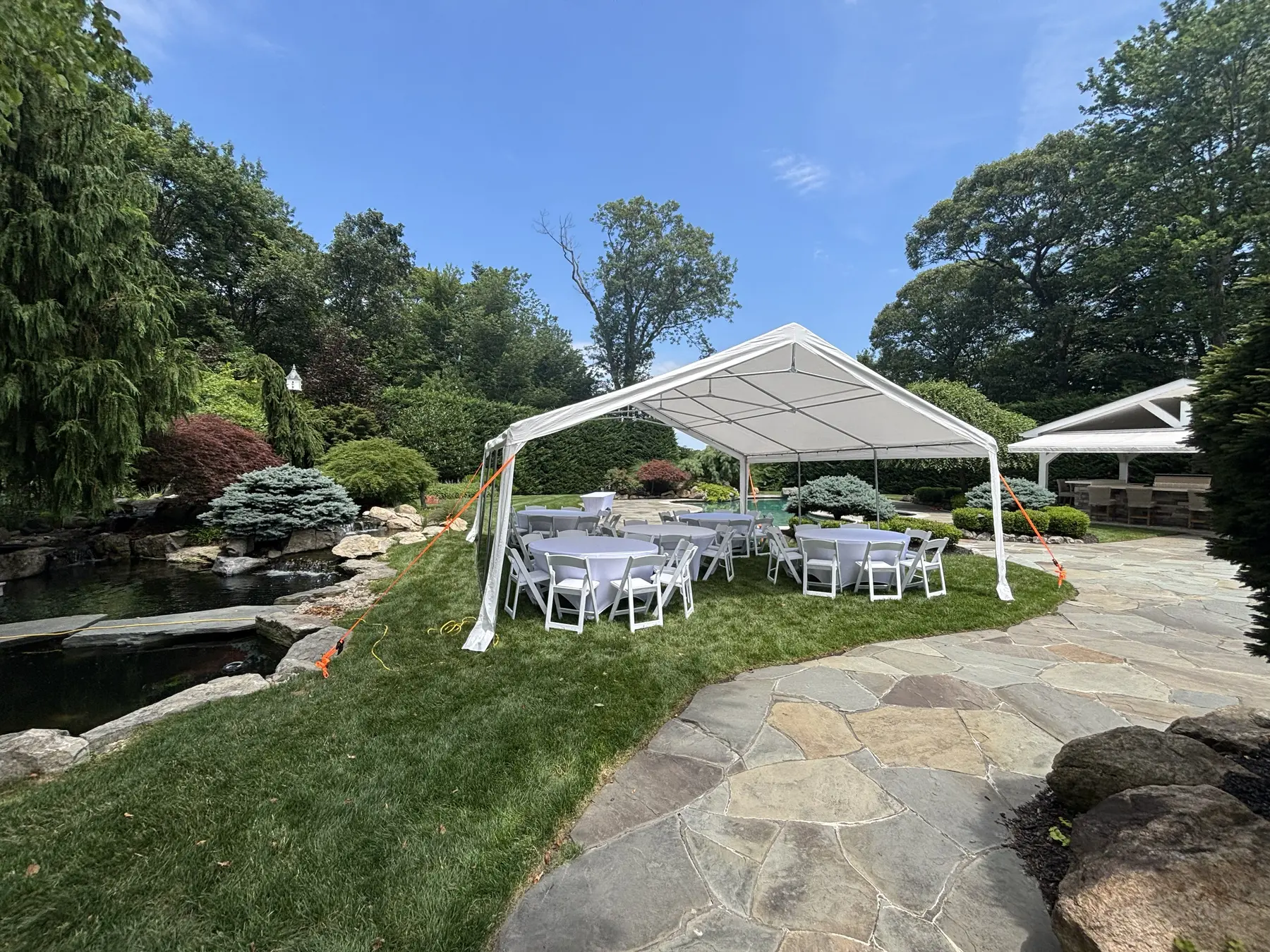 White frame tent with round tables set up beside a landscaped backyard pond and pool