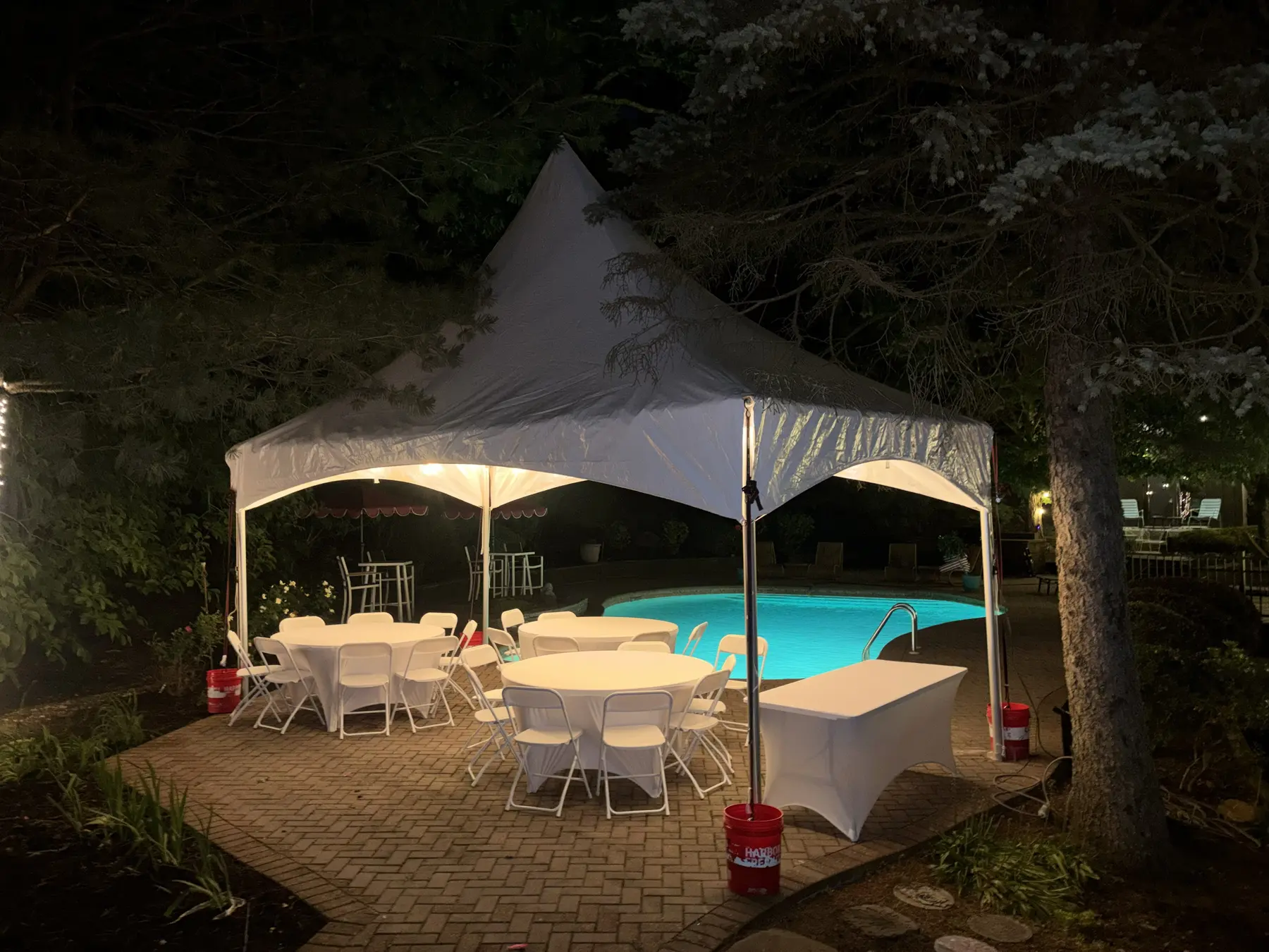 Illuminated high-peak tent with round tables and a cocktail table beside a backyard pool